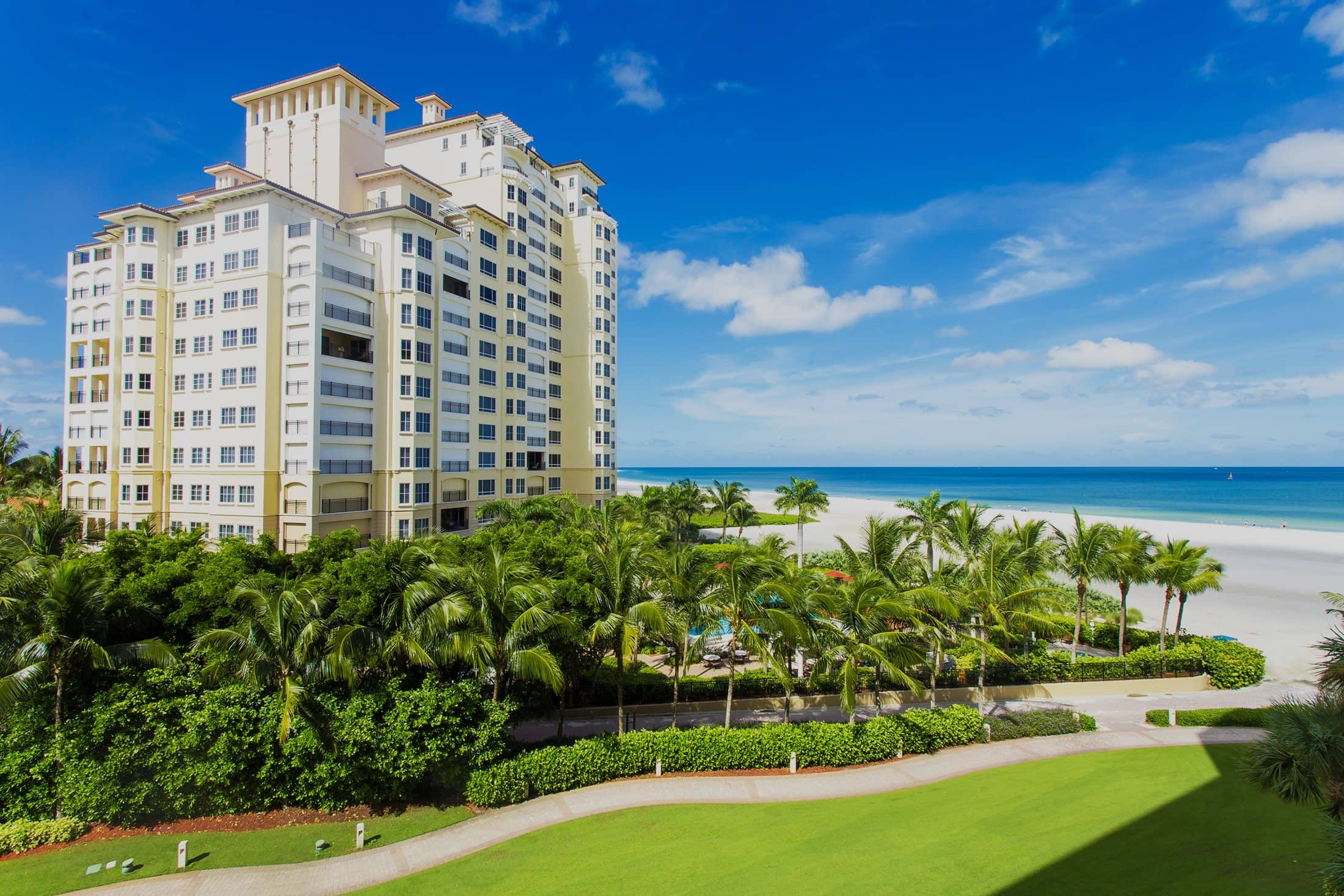 Coastal luxury condominium tower beside palm gardens and a white-sand beach